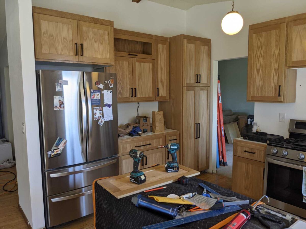 remodel of kitchen cabinetry installation in process for turning makeshift office space on room dividing wall into storage rich, compact kitchen space, with room to hold a full size french door refrigerator with bottom drawers. White oak cabinetry with general finish to reveal the natural beauty of white oak wood